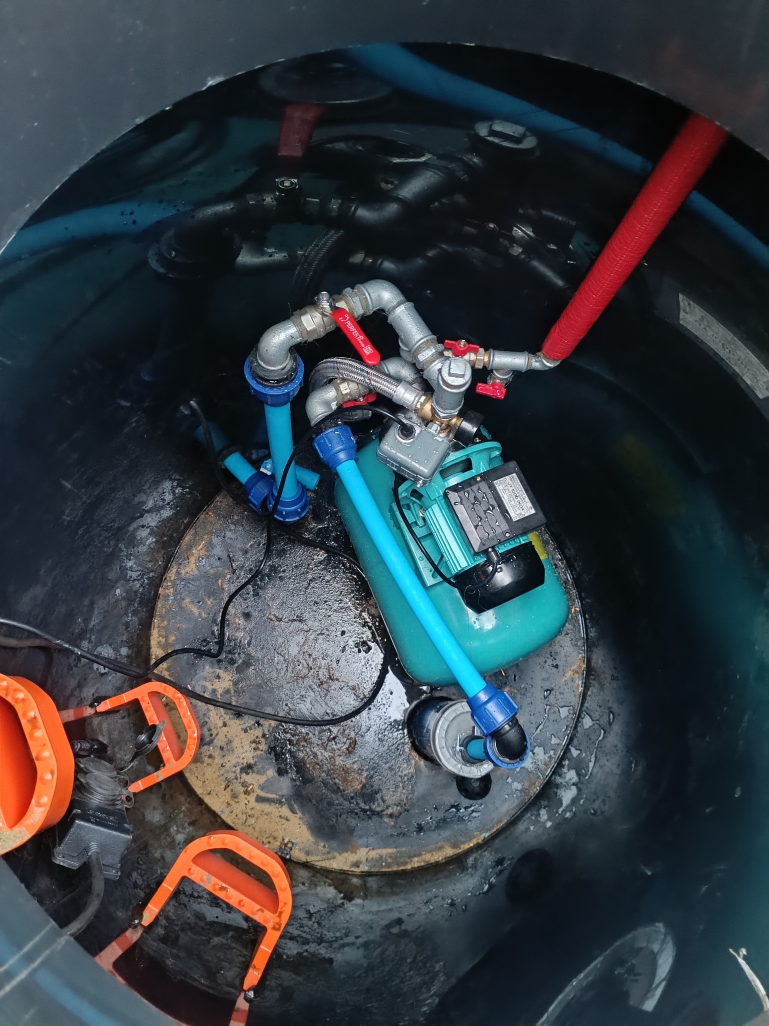 Hydrofor w studni: zielony zbiornik, niebieskie rury, metalowe zawory z czerwonymi uchwytami. Widoczne pomarańczowe uchwyty i czarne kable. Wnętrze studni z wilgocią.