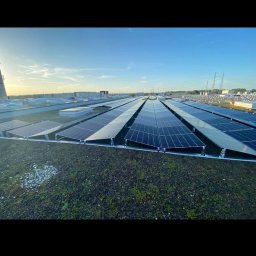 Elektro-Flesz Fotowoltaika oraz Magazyn Energi - Rozległa instalacja fotowoltaiczna na zielonym dachu budynku, widok z góry, błękitne niebo w tle. Panele ułożone rzędami, równomierne oświetlenie.