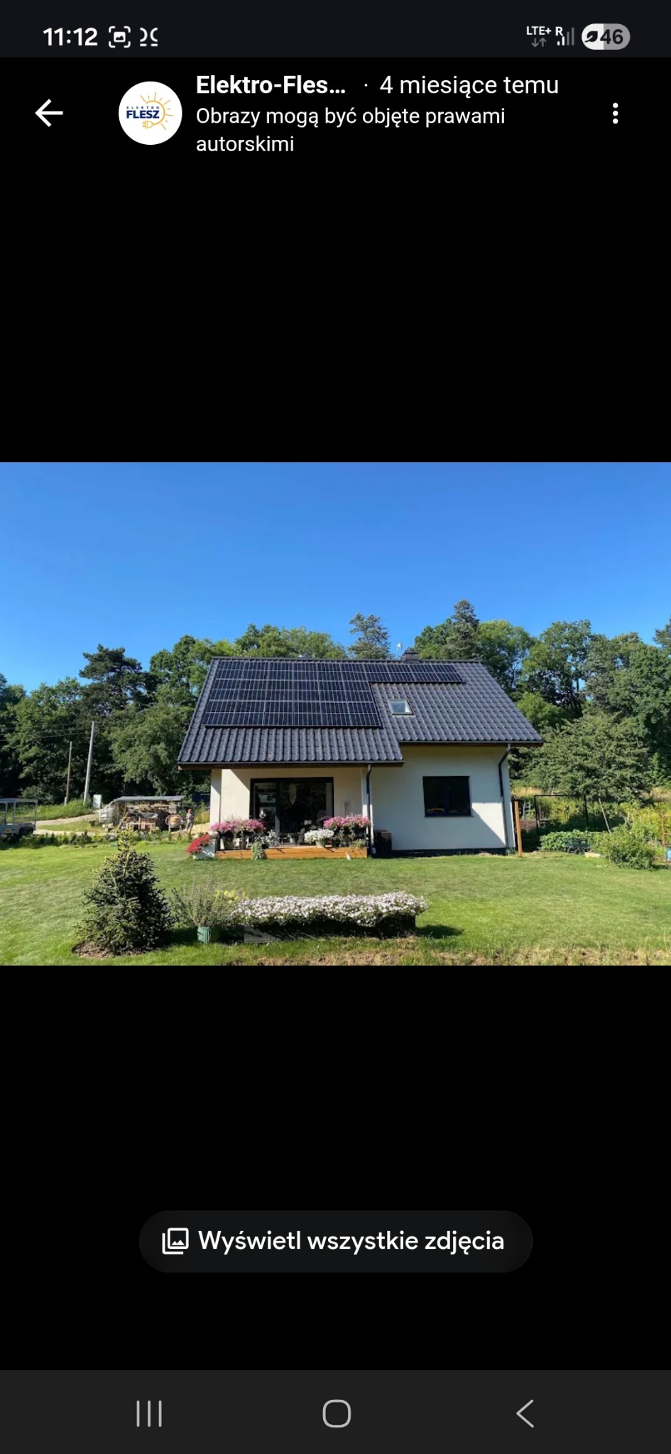 Dom jednorodzinny z panelami słonecznymi na dachu, otoczony zielonym trawnikiem i kwiatami. Ujęcie z poziomu ogrodu, w słoneczny dzień.