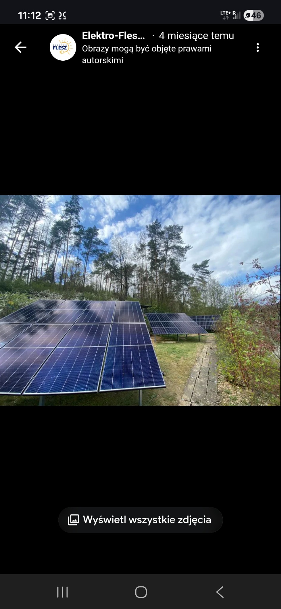 Instalacja paneli na trawiastym terenie, widok z dołu na niebo i drzewa. Panele ustawione rzędami, widoczna ścieżka z płyt. Ujęcie z niskiej perspektywy.