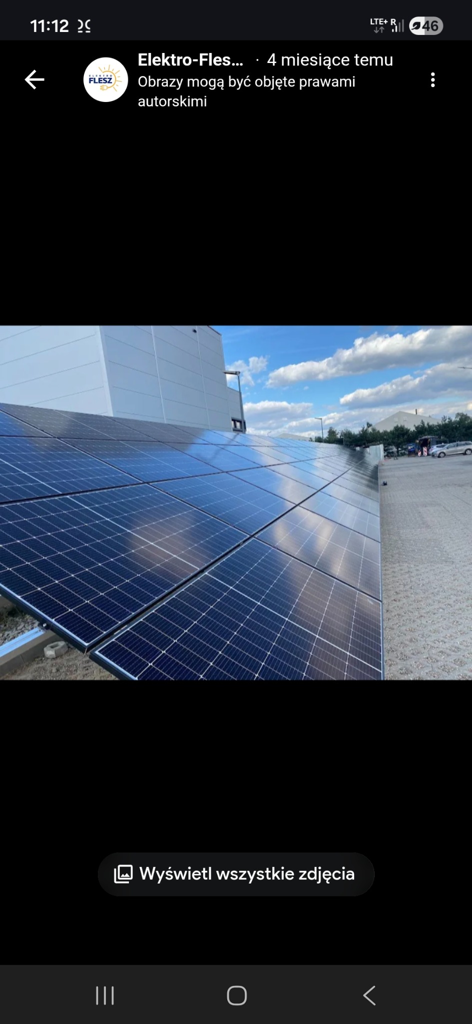 Błękitne panele na dachu budynku przemysłowego odbijają niebo. W tle zaparkowane samochody i drzewa. Widok z dołu, lekko pod kątem.