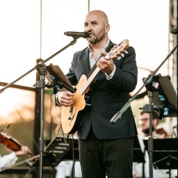 Marcin Pomykała - tenor z gitarą - Wokalista z gitarą na scenie, w tle orkiestra smyczkowa. Mężczyzna w garniturze śpiewa do mikrofonu, grając na gitarze akustycznej. Zespół muzyczny podczas występu.
