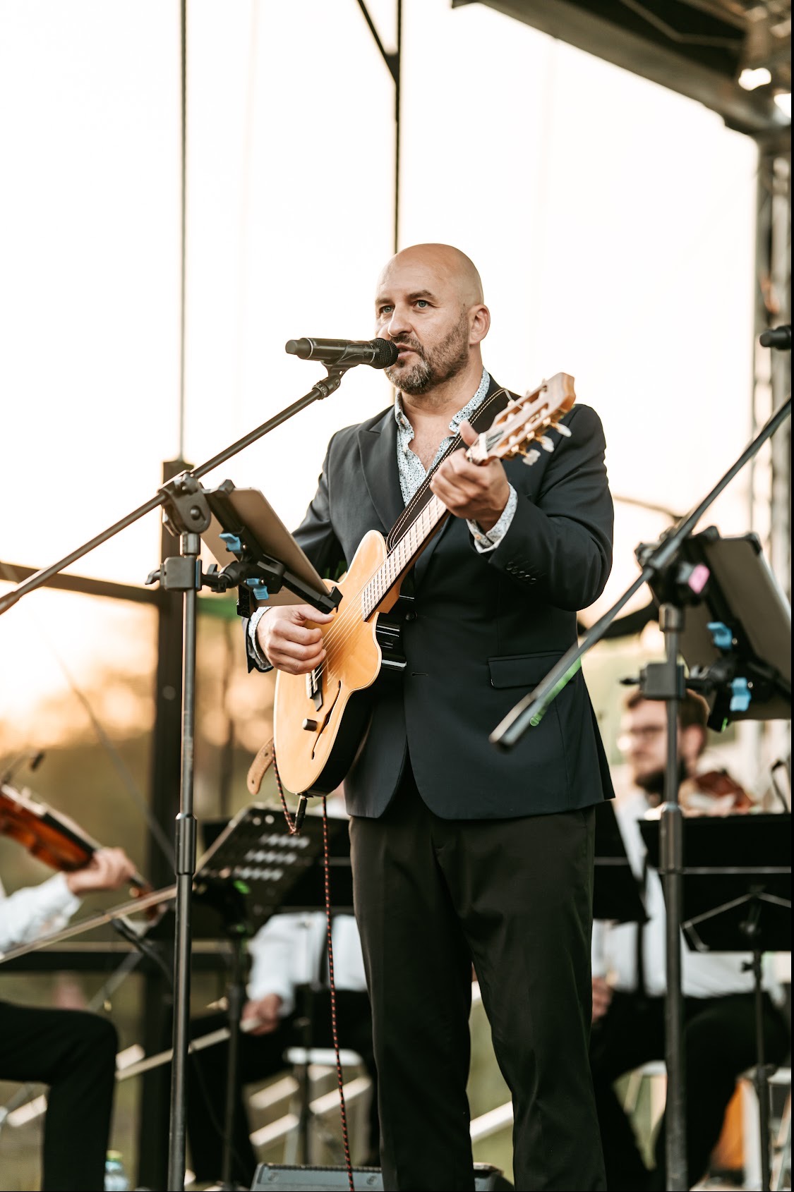 Wokalista z gitarą na scenie, w tle orkiestra smyczkowa. Mężczyzna w garniturze śpiewa do mikrofonu, grając na gitarze akustycznej. Zespół muzyczny podczas występu.