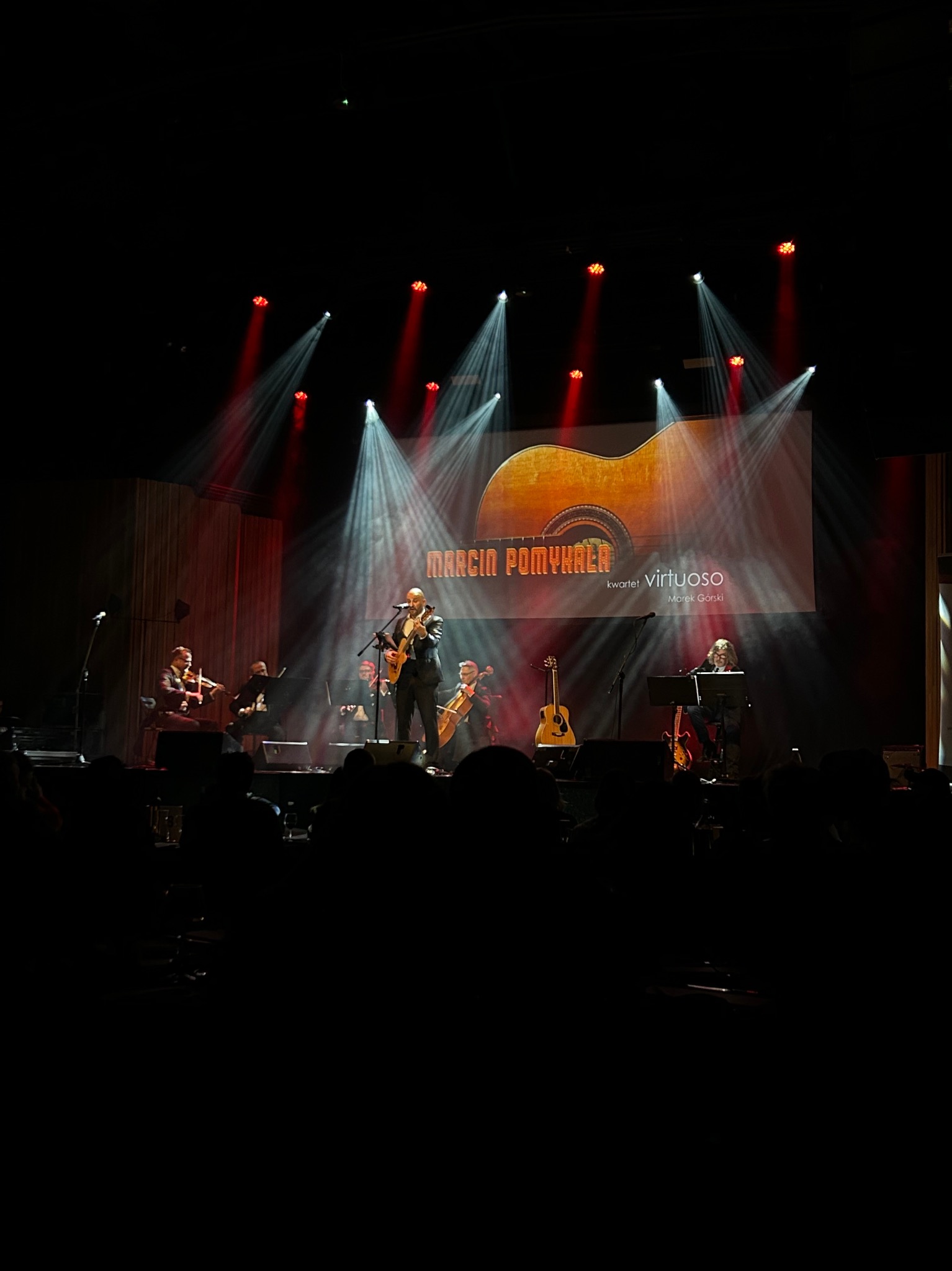 Zespół muzyczny na scenie podczas koncertu. Wokalista z gitarą, skrzypce, wiolonczela, oświetlenie sceniczne i publiczność w tle. Marcin Pomykała Virtuoso.