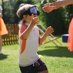 Lexy Event Agency - Chłopiec w okularach do nurkowania, w ruchu na trawie, wyciąga ręce do osoby w pomarańczowej sukience. Animacje plenerowe w słoneczny dzień.