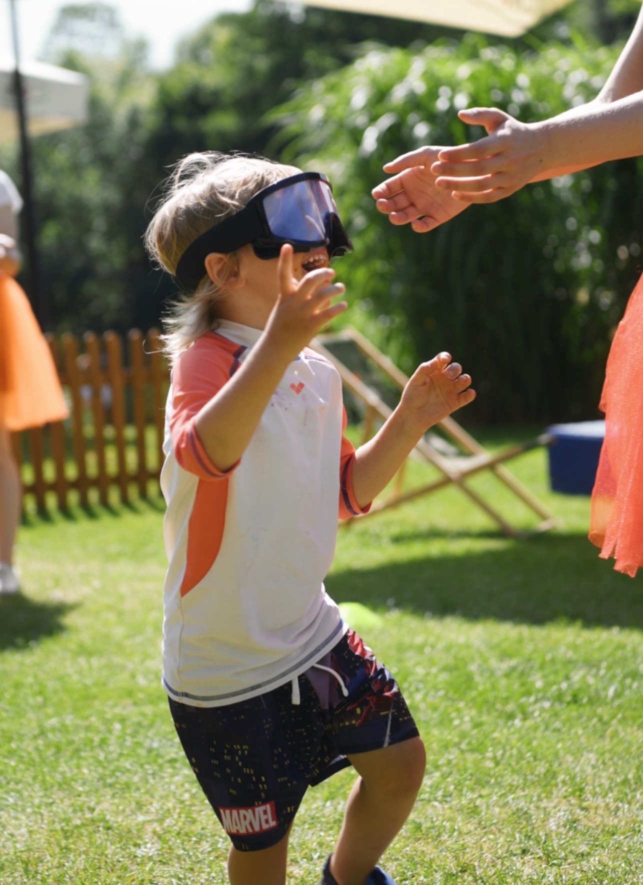 Chłopiec w okularach do nurkowania, w ruchu na trawie, wyciąga ręce do osoby w pomarańczowej sukience. Animacje plenerowe w słoneczny dzień.