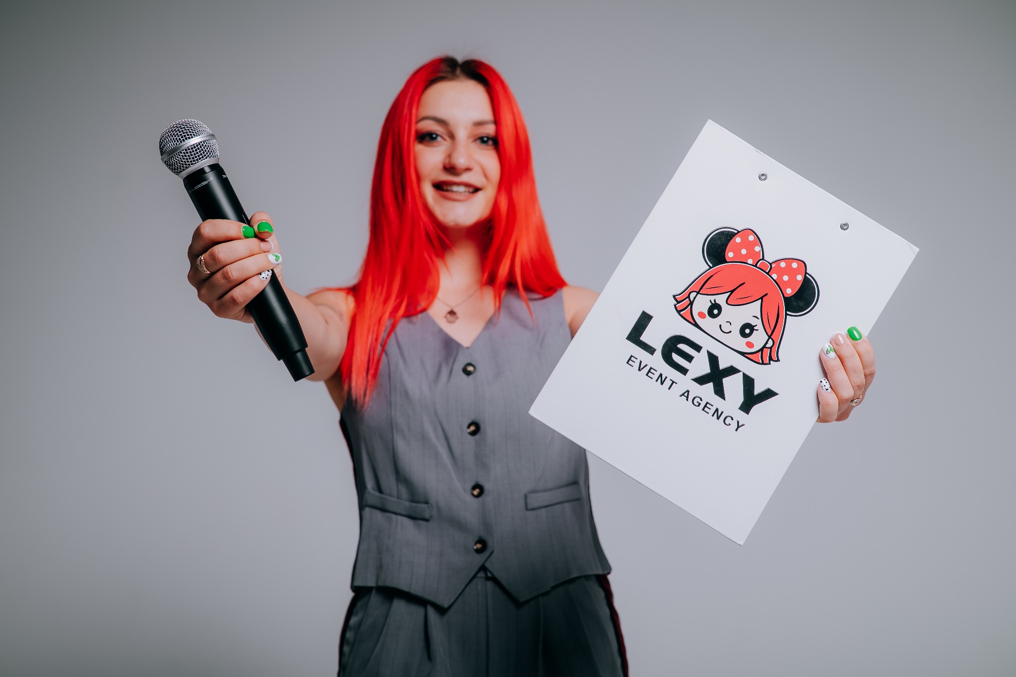 Uśmiechnięta kobieta z czerwonymi włosami prezentuje mikrofon i logo Lexy Event Agency na białej podkładce, na neutralnym tle. Widoczne detale manicure.