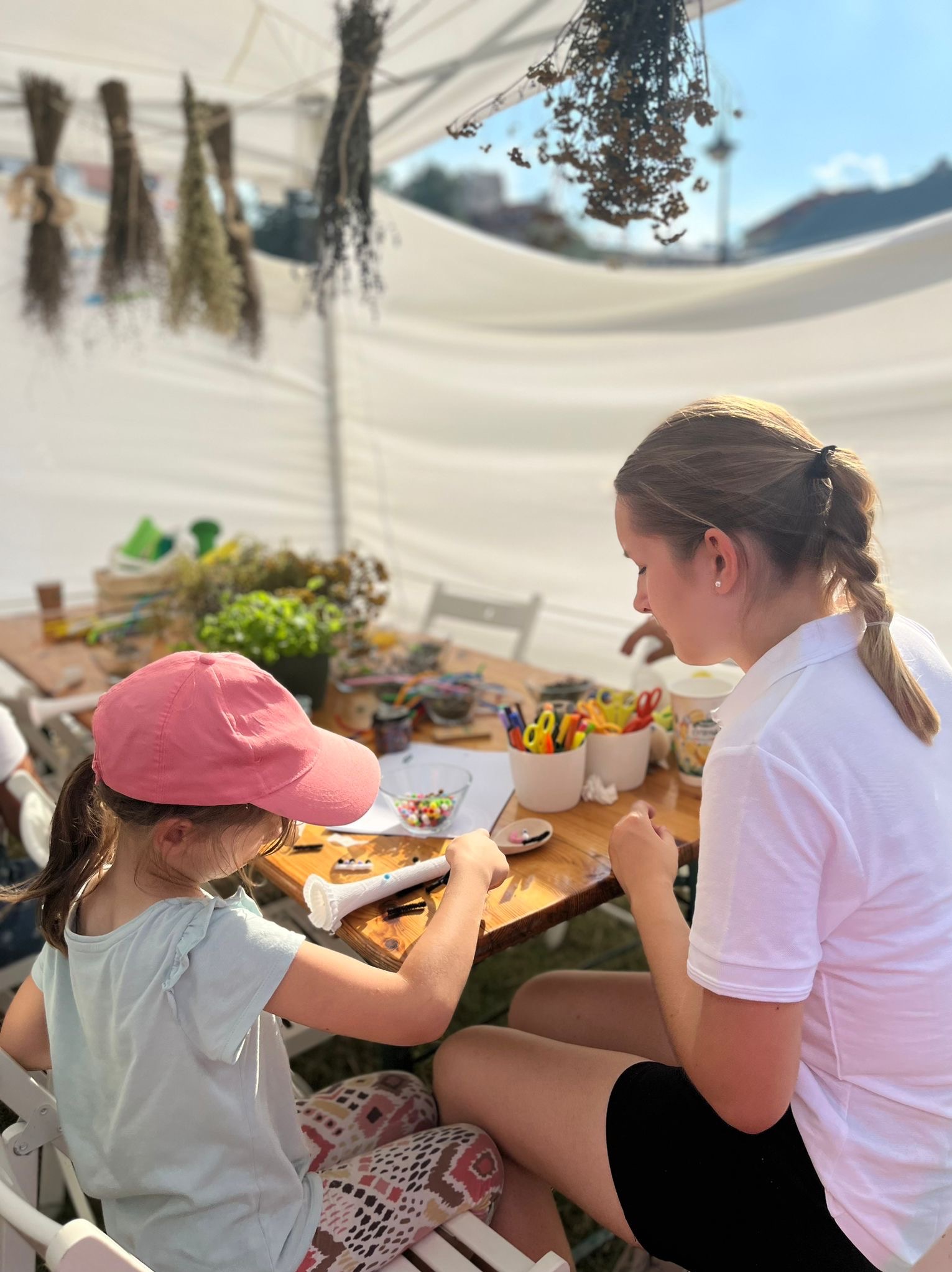 Warsztaty ekologiczne: dziewczynka w różowej czapce i kobieta tworzą ozdoby z naturalnych materiałów przy drewnianym stole pod białym namiotem.