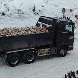 TomTrack Tomasz Sobierajski - Czarna wywrotka Scania wypełniona pociętym drewnem na tle zaśnieżonego krajobrazu. Widoczne drzewa i krzewy pokryte śniegiem. Zima.