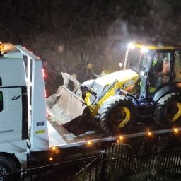 TomTrack Tomasz Sobierajski - Żółta koparko-ładowarka JCB transportowana na białej lawecie MAN TGS 26.440 w nocy. Widoczny fragment zbocza i ogrodzenie.
