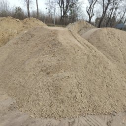 TomTrack Tomasz Sobierajski - Góra piasku z widocznymi śladami opon, w tle drzewa bez liści. Piasek o jasnym, beżowym odcieniu. Widoczne grudki i nierówności na powierzchni.