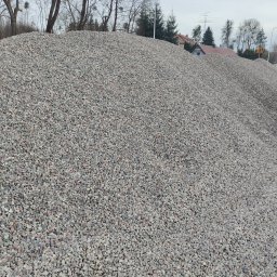 TomTrack Tomasz Sobierajski - Góra szarego kruszywa na tle zabudowań i drzew. Widoczna faktura kamieni i ich zróżnicowana wielkość. Kruszywo tworzy dużą pryzmę.