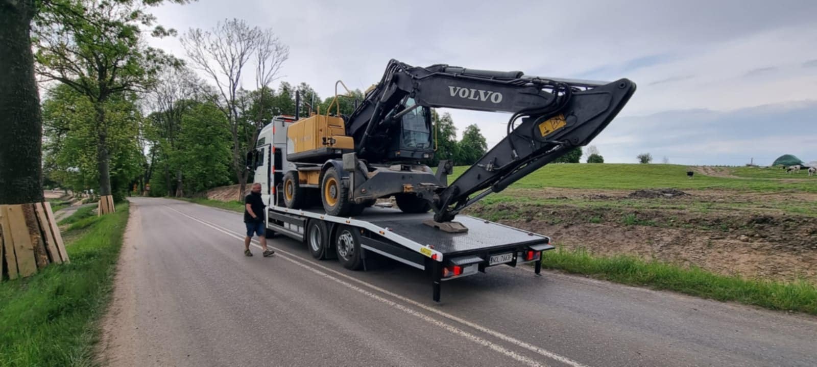 Laweta przewozi czarno-żółtą koparkę Volvo na tle zielonego krajobrazu. Mężczyzna w czarnej koszulce idzie obok lawety po asfaltowej drodze.