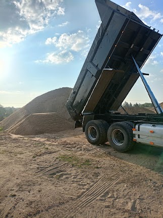 Ciężarówka z podniesioną skrzynią wyładowuje piasek na placu budowy w słoneczny dzień. Widoczne ślady opon na ziemi i kopiec piasku.