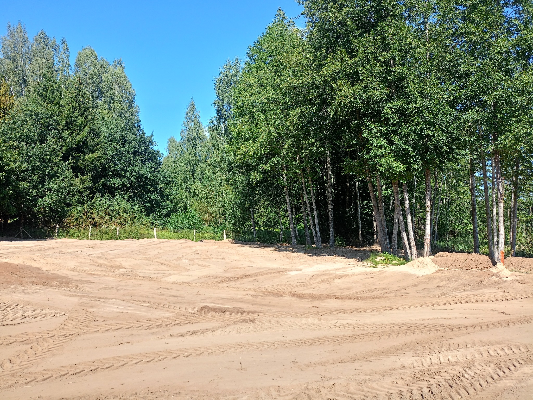 Wyrównany teren pod budowę z widocznymi śladami opon, otoczony zielenią drzew i błękitnym niebem. Przygotowany grunt pod inwestycję.
