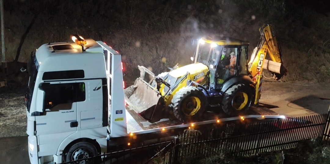 Żółta koparko-ładowarka JCB transportowana na białej lawecie MAN TGS 26.440 w nocy. Widoczny fragment zbocza i ogrodzenie.