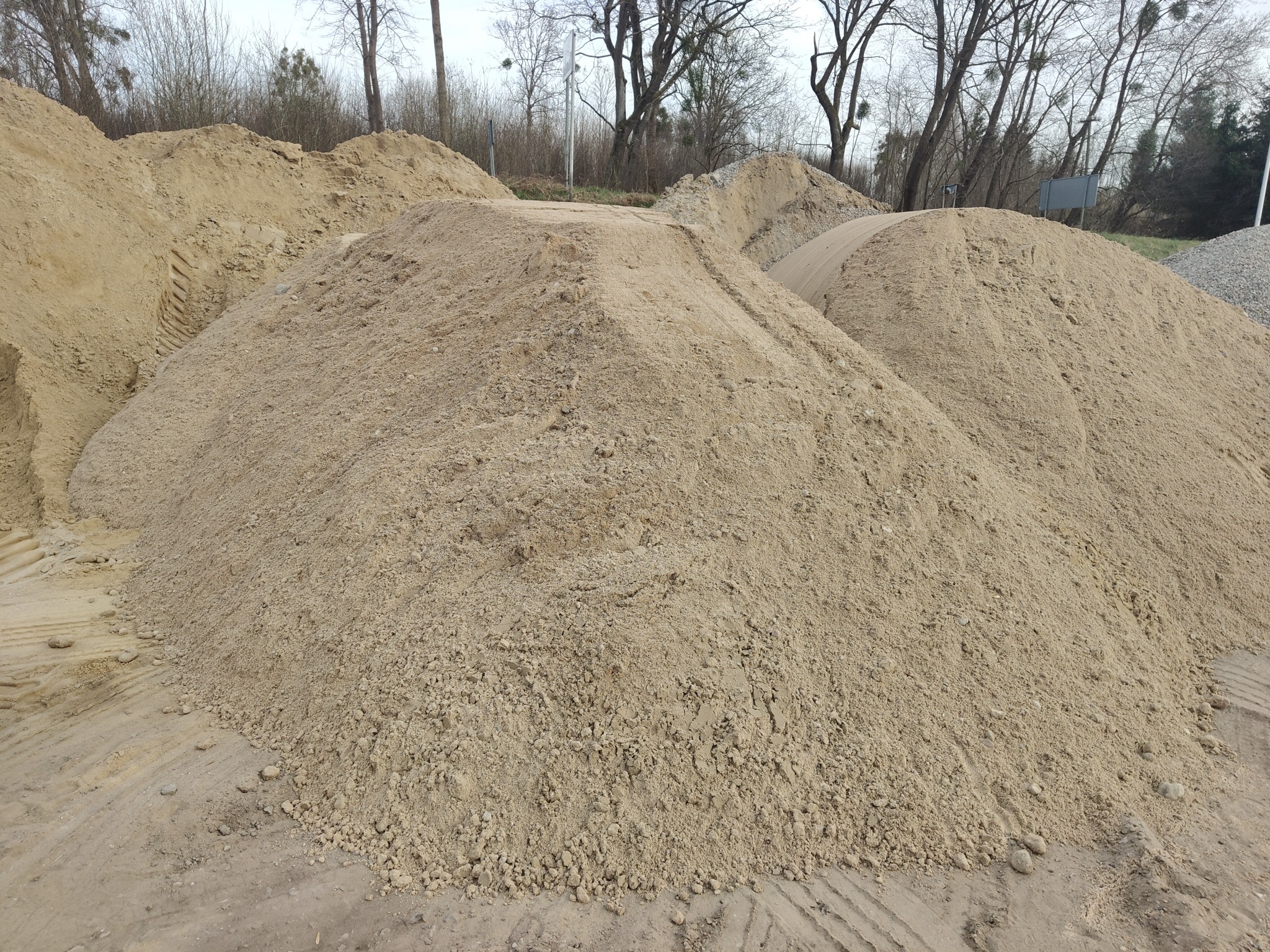 Góra piasku z widocznymi śladami opon, w tle drzewa bez liści. Piasek o jasnym, beżowym odcieniu. Widoczne grudki i nierówności na powierzchni.