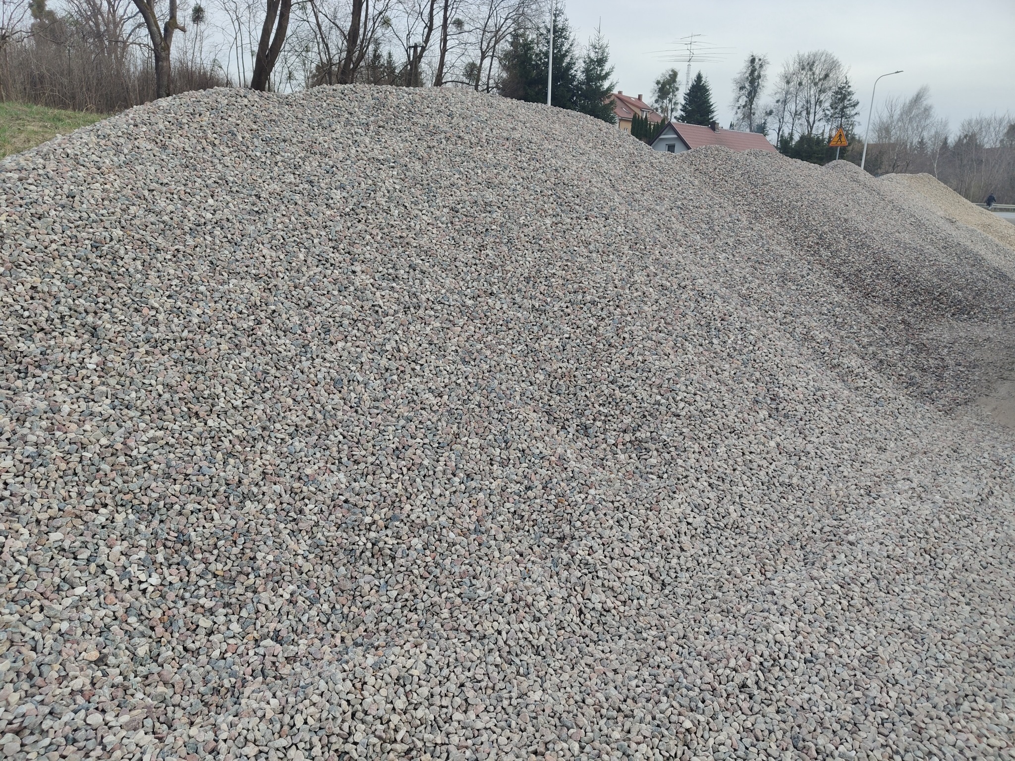 Góra szarego kruszywa na tle zabudowań i drzew. Widoczna faktura kamieni i ich zróżnicowana wielkość. Kruszywo tworzy dużą pryzmę.