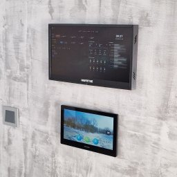 Home Koncept Jarosław Dukat - Dwa panele dotykowe na ścianie o fakturze betonu: górny z interfejsem inteligentnego domu, dolny z podglądem z kamery. Widoczny fragment drzwi z czarną ramą i włącznik.