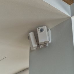 Home Koncept Jarosław Dukat - Dwie białe kamery IP zamontowane w rogu pomieszczenia, widoczne diody sygnalizacyjne. Minimalistyczny design, szara ściana i biały sufit. Ujęcie z dołu.