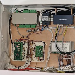 Home Koncept Jarosław Dukat - Otwarta szafka z zamontowanymi modułami sterującymi, przewodami i zasilaczem. Widoczny moduł Hikvision i inne komponenty. Klucze do szafki wiszą po lewej stronie.