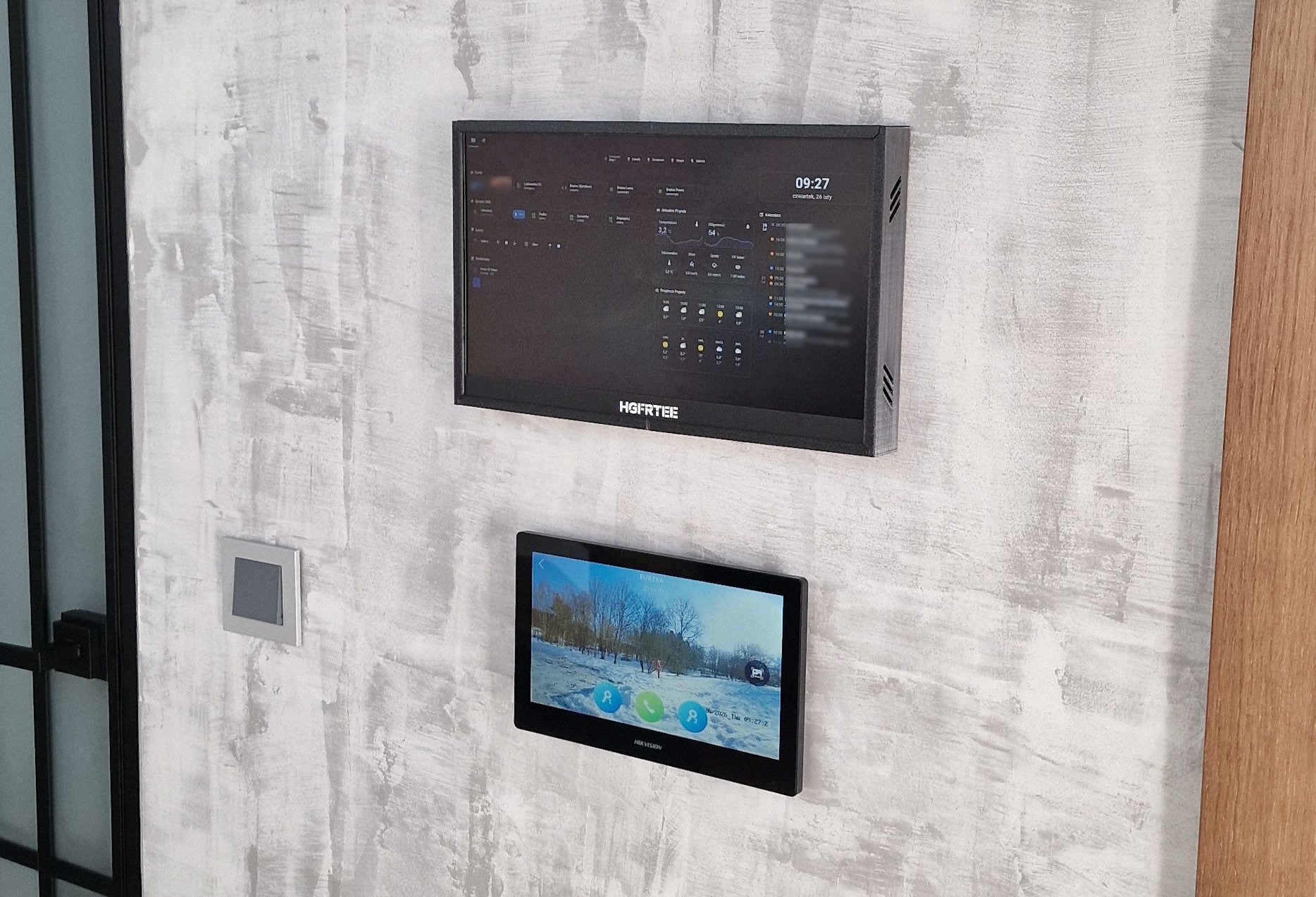 Dwa panele dotykowe na ścianie o fakturze betonu: górny z interfejsem inteligentnego domu, dolny z podglądem z kamery. Widoczny fragment drzwi z czarną ramą i włącznik.