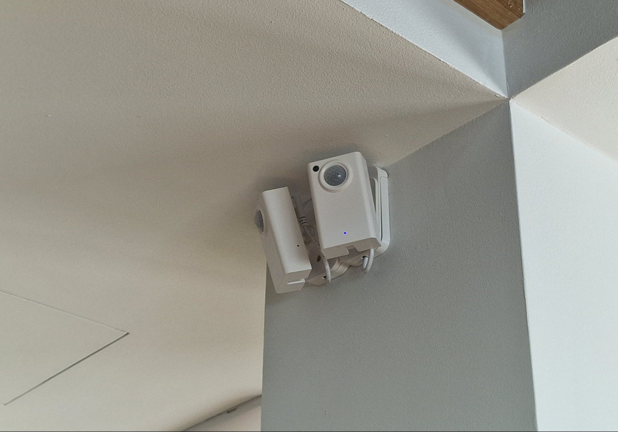 Dwie białe kamery IP zamontowane w rogu pomieszczenia, widoczne diody sygnalizacyjne. Minimalistyczny design, szara ściana i biały sufit. Ujęcie z dołu.