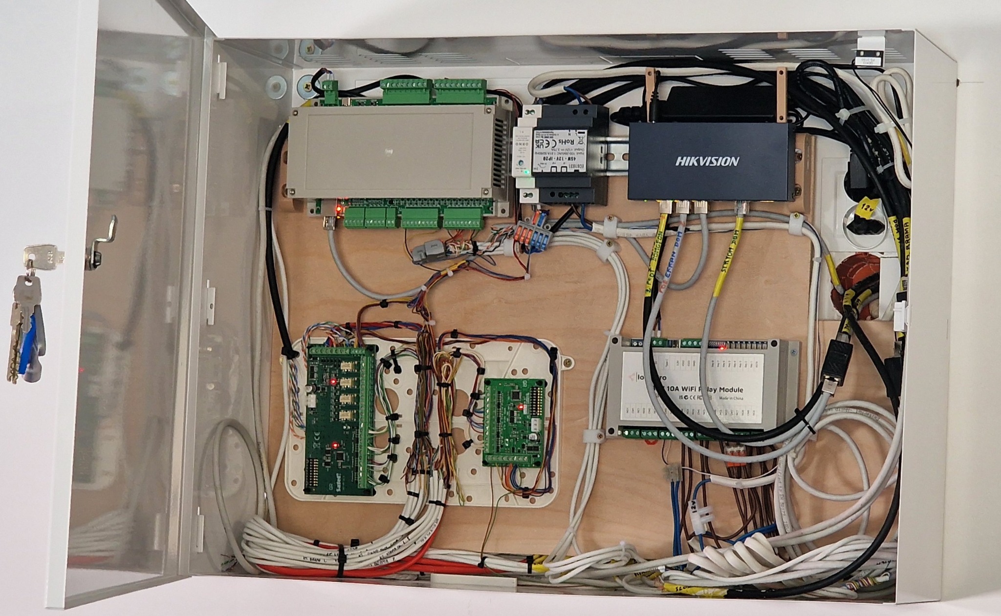 Otwarta szafka z zamontowanymi modułami sterującymi, przewodami i zasilaczem. Widoczny moduł Hikvision i inne komponenty. Klucze do szafki wiszą po lewej stronie.