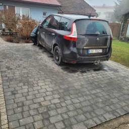 Marcin Maj - Szara kostka brukowa na podjeździe przed domem, zaparkowany czarny samochód Renault Scenic. Widoczne budynki w tle i elementy zieleni.