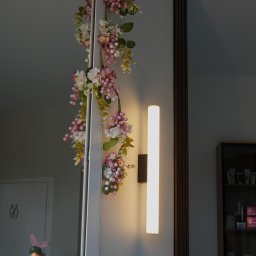 Bloom Skin Sp Z O.O. - Klimatyczne wnętrze: dekoracja kwiatowa przy lustrze, pionowa lampa ścienna i regał z kosmetykami w tle. Przytulna atmosfera w gabinecie.