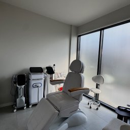 Bloom Skin Sp Z O.O. - Jasne wnętrze gabinetu z nowoczesnym fotelem zabiegowym Calissimo, sprzętem kosmetycznym i krzesłem na kółkach przy oknie z mleczną szybą.