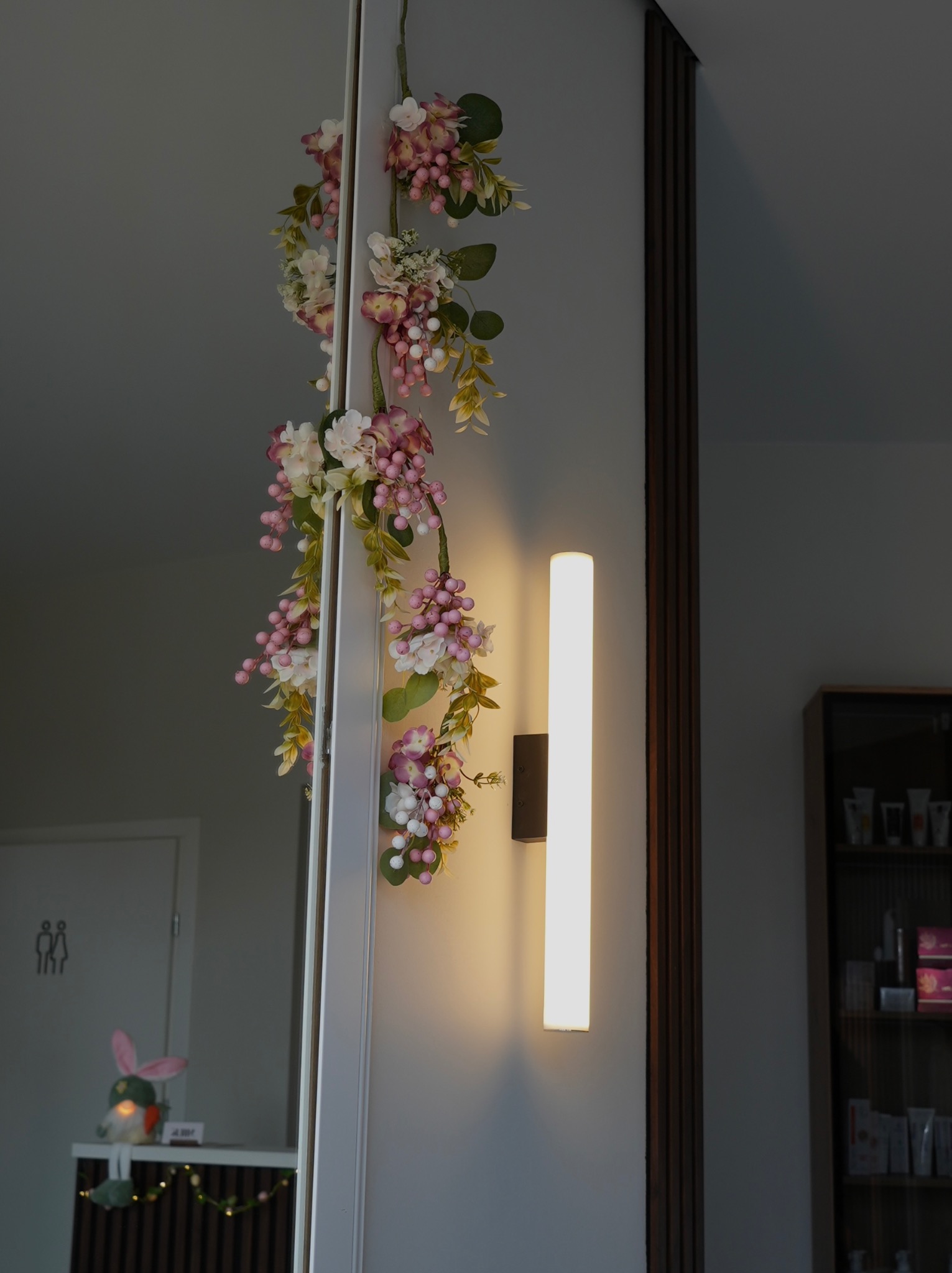 Klimatyczne wnętrze: dekoracja kwiatowa przy lustrze, pionowa lampa ścienna i regał z kosmetykami w tle. Przytulna atmosfera w gabinecie.