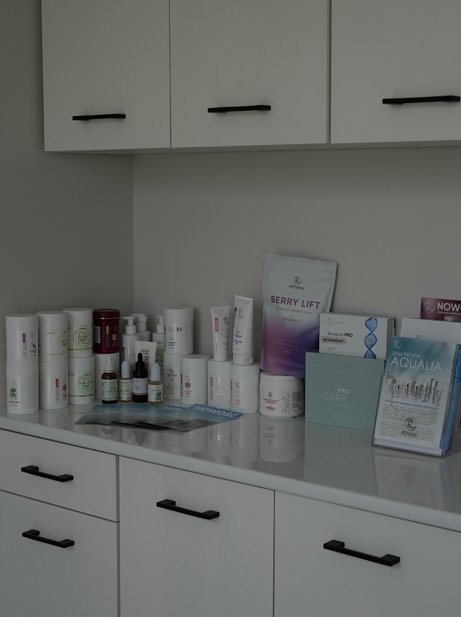 Różnorodne produkty marki Renew na białej szafce z czarnymi uchwytami, w tle białe szafki wiszące. Kosmetyki do pielęgnacji twarzy i ciała w gabinecie.