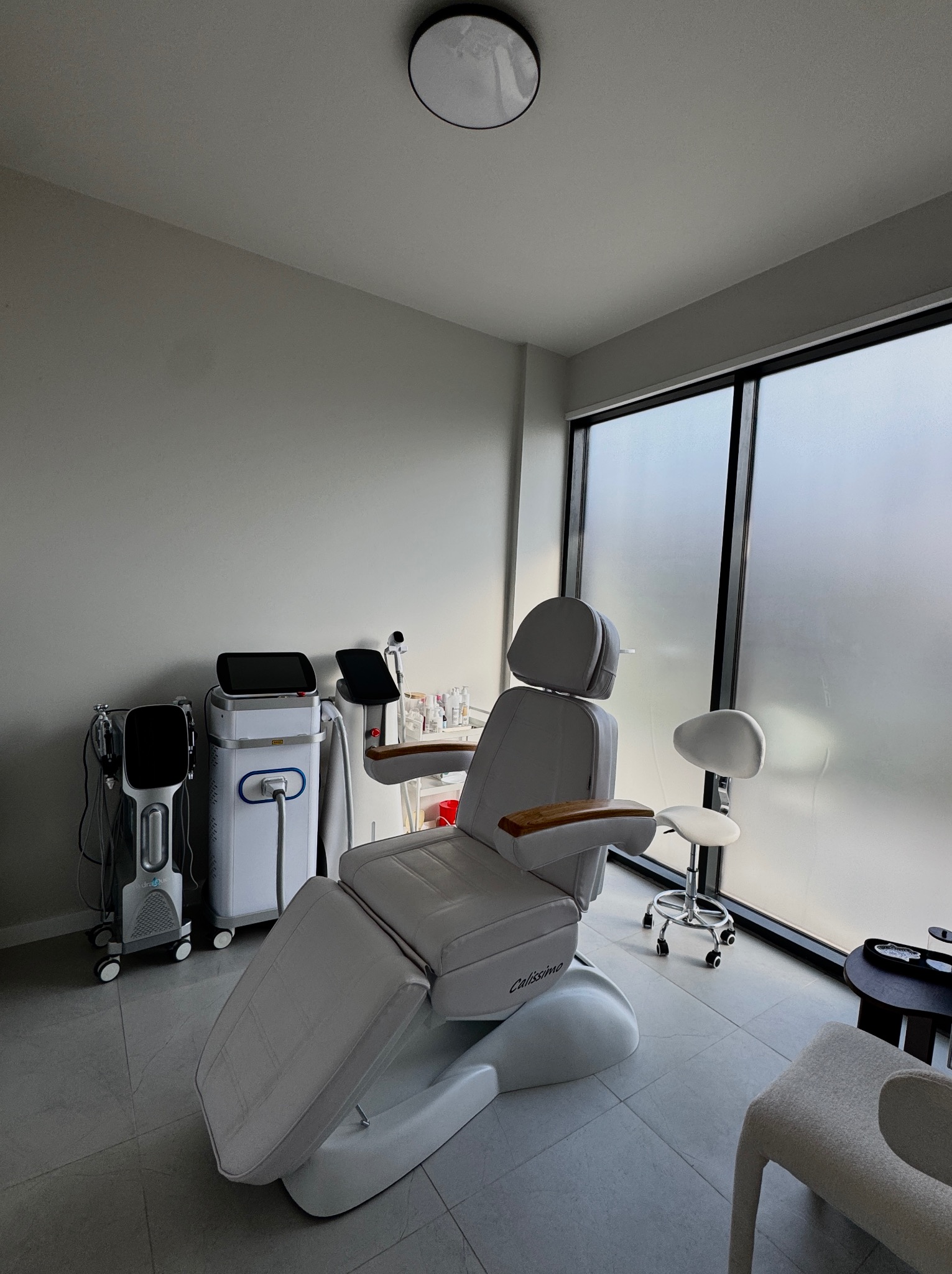 Jasne wnętrze gabinetu z nowoczesnym fotelem zabiegowym Calissimo, sprzętem kosmetycznym i krzesłem na kółkach przy oknie z mleczną szybą.