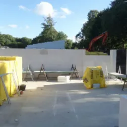 Budowa domu w trakcie: wznoszone ściany z betonu komórkowego, widoczne materiały budowlane w żółtych opakowaniach, koparka w tle.