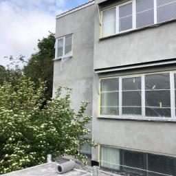 Adeco - Dwu-kondygnacyjny budynek w trakcie szpachlowania elewacji, okna zabezpieczone folią i taśmą, na pierwszym planie rusztowanie z wałkiem malarskim.