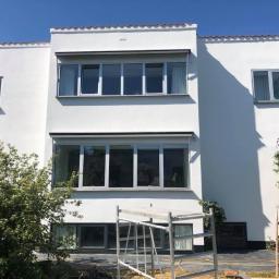 Adeco - Dwukondygnacyjny budynek o białej, gładkiej elewacji w stylu modernistycznym, z dużymi oknami, otoczony zielenią i z rozstawionym rusztowaniem malarskim na pierwszym planie.