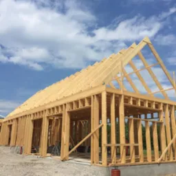 Konstrukcja szkieletowa domu w trakcie budowy, widoczne drewniane belki, słupy i krokwie na fundamencie, z widokiem na błękitne niebo z chmurami w tle.