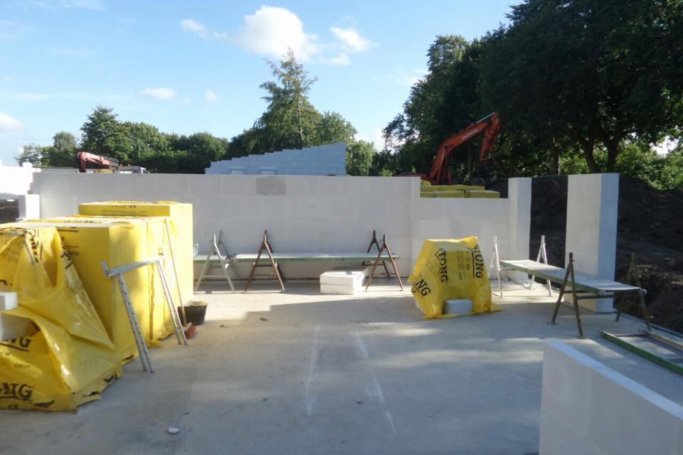 Budowa domu w trakcie: wznoszone ściany z betonu komórkowego, widoczne materiały budowlane w żółtych opakowaniach, koparka w tle.