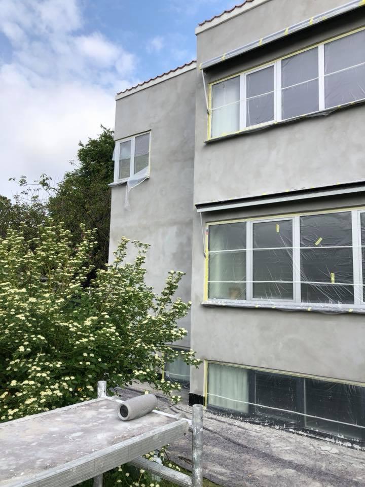 Dwu-kondygnacyjny budynek w trakcie szpachlowania elewacji, okna zabezpieczone folią i taśmą, na pierwszym planie rusztowanie z wałkiem malarskim.