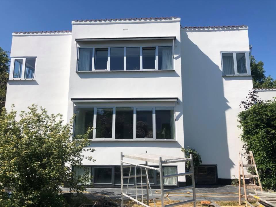 Dwukondygnacyjny budynek o białej, gładkiej elewacji w stylu modernistycznym, z dużymi oknami, otoczony zielenią i z rozstawionym rusztowaniem malarskim na pierwszym planie.