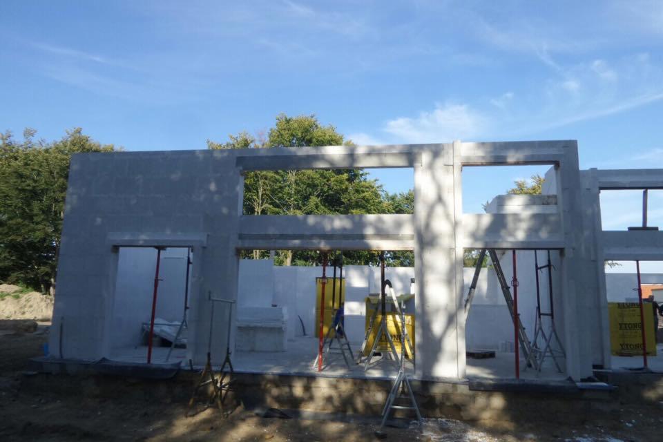 Surowa konstrukcja budynku z betonu komórkowego, widoczne otwory okienne i drzwiowe, wsparta metalowymi stemplami i drabinkami na placu budowy z drzewami w tle.