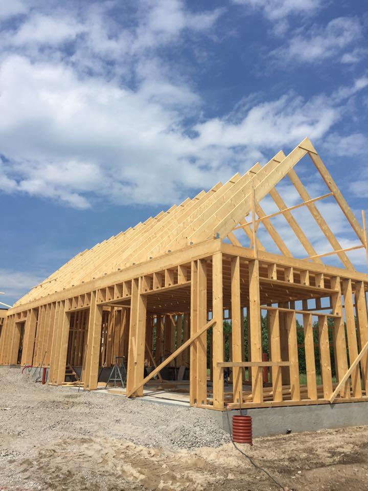 Konstrukcja szkieletowa domu w trakcie budowy, widoczne drewniane belki, słupy i krokwie na fundamencie, z widokiem na błękitne niebo z chmurami w tle.