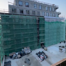 Jan Ojczyk Usługi Budowlane - Budynek w trakcie budowy, osłonięty zieloną siatką rusztowaniową. Widoczne okna i elementy konstrukcyjne. Na dole materiały budowlane i teren budowy.