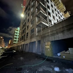 Jan Ojczyk Usługi Budowlane - Nocne ujęcie surowego budynku w trakcie budowy, z widocznym żurawiem i materiałami budowlanymi. Oświetlenie uliczne podkreśla betonową konstrukcję.