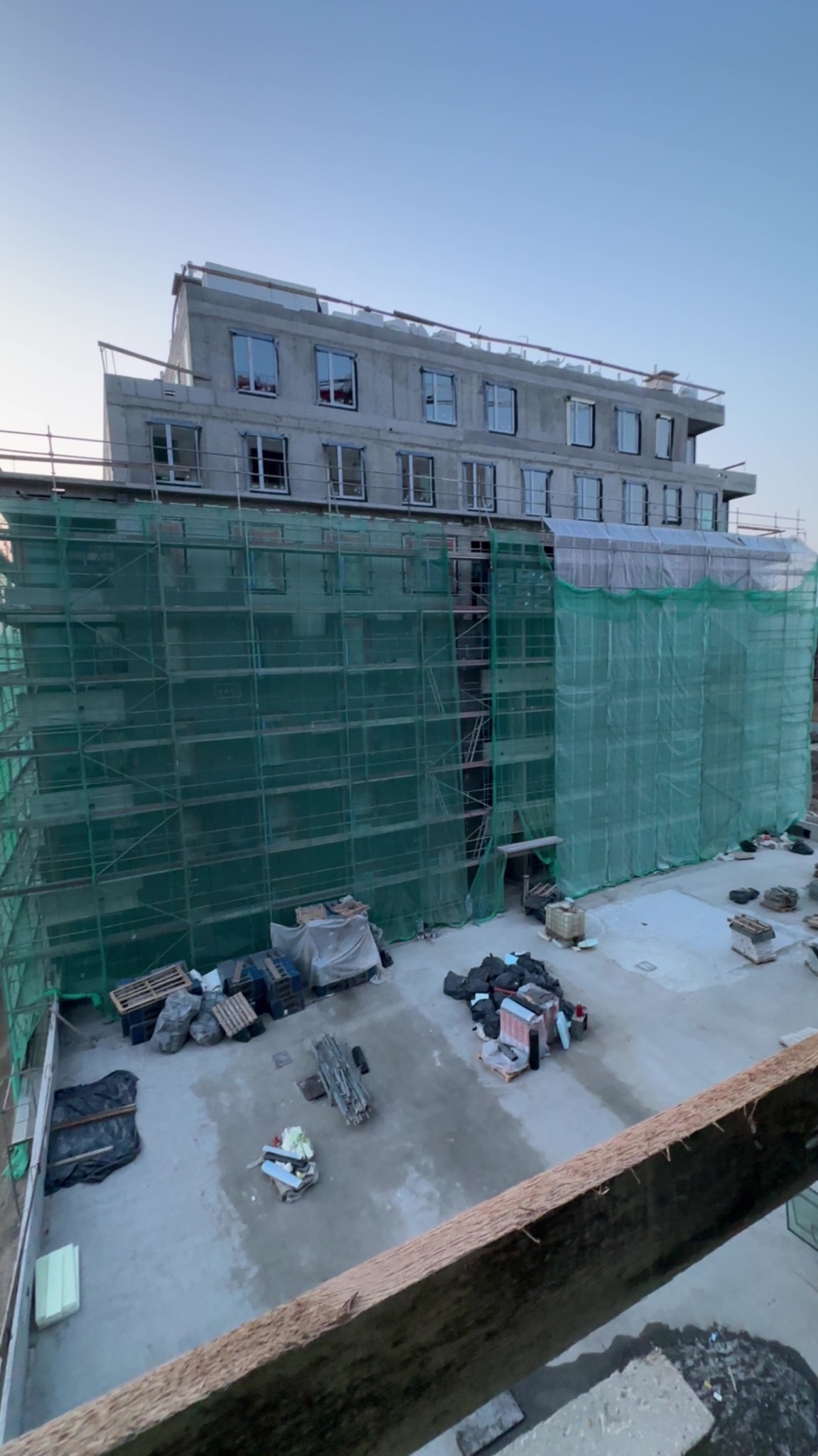 Budynek w trakcie budowy, osłonięty zieloną siatką rusztowaniową. Widoczne okna i elementy konstrukcyjne. Na dole materiały budowlane i teren budowy.