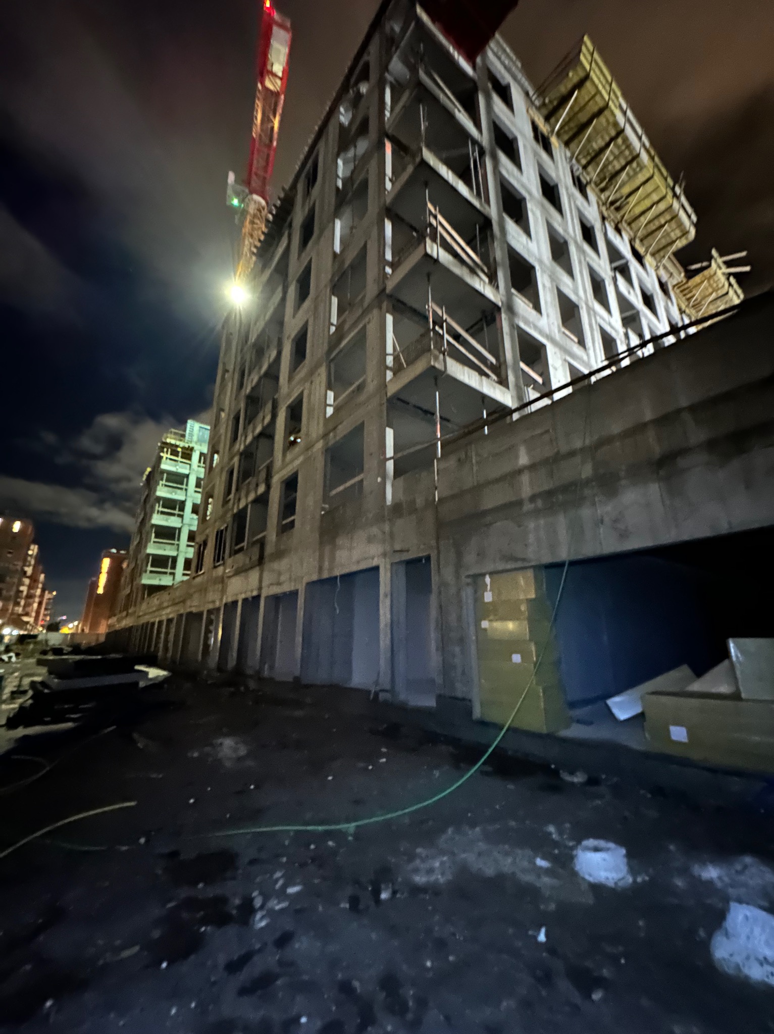 Nocne ujęcie surowego budynku w trakcie budowy, z widocznym żurawiem i materiałami budowlanymi. Oświetlenie uliczne podkreśla betonową konstrukcję.