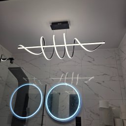 Mateusz Myśków Usługi Elektryczne - Nowoczesna lampa LED w kształcie spirali, zamontowana w łazience z marmurowymi płytkami i okrągłymi lustrami z podświetleniem. Widoczna kabina prysznicowa.