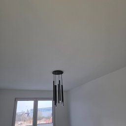 Mateusz Myśków Usługi Elektryczne - Czarny, nowoczesny żyrandol z podłużnymi lampami w Wałbrzychu, zamontowany na białym suficie. W tle okno z widokiem na zewnątrz i fragment czarnej sofy.