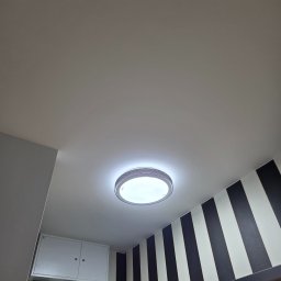 Mateusz Myśków Usługi Elektryczne - Zamontowana okrągła lampa sufitowa LED w łazience z czarno-białymi pasami na ścianie. Widok z dołu, oświetlenie jasne, nowoczesny design.